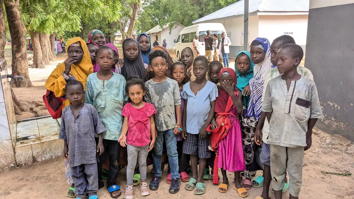 Women and children seated on the ground outdoors at a medical outreach.